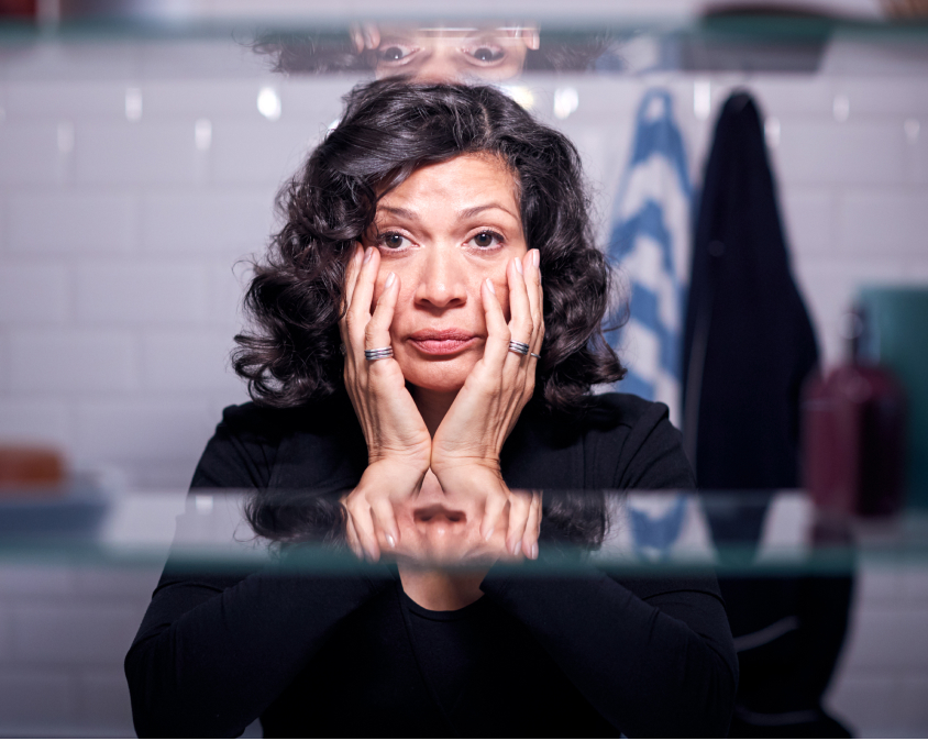 Woman looking into a bathroom mirror with her hands on her face