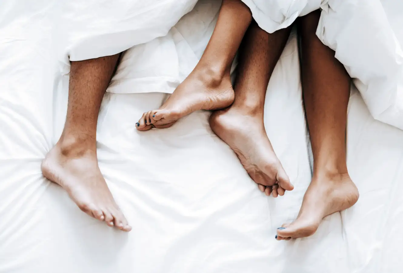 Two pairs of feet sticking out from underneath a white sheet on a white bed